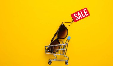 Alışveriş arabası ve sarı arka planda kırmızı etiketli şık güneş gözlükleri. Üst Manzara. İndirim. Minimalizm