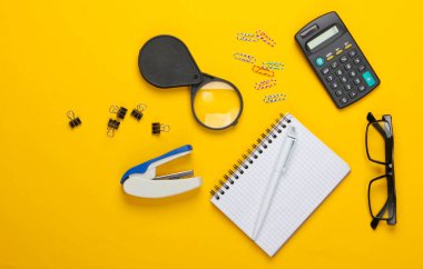 Ofis aletleri, kırtasiye malzemeleri. Zımba, hesap makinesi, büyüteç, ataç, sarı arka planda defter. Üst görünüm