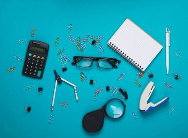 Ofis aletleri, kırtasiye malzemeleri. Zımba, hesap makinesi, büyüteç, ataç, not defteri, mavi arka planda pusula. Üst görünüm