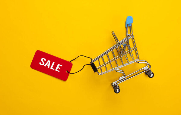 Mini shopping trolley with sale tag on yellow background. Top view. Discount. Minimalism