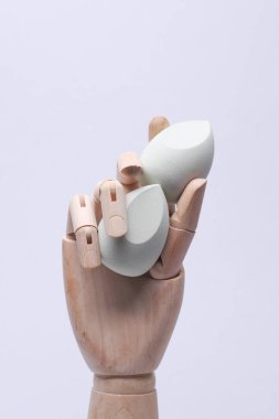 Kuklanın tahta eli gri arka planda makyaj süngeri tutuyor.