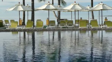 Yüzme havuzu manzarası, plaj şemsiyeleri, palmiye ağaçları ve mavi gökyüzü.