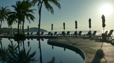 Yüzme Havuzu, plaj şemsiyeleri, palmiye ağaçları ve dramatik gökyüzü günbatımı manzarası
