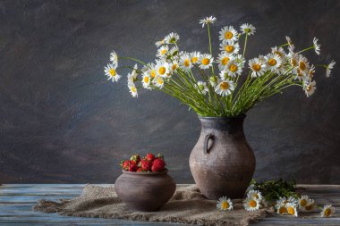 Natürmort. Bir demet chamomiles, çilek ve tahta bir masanın üzerinde kil sürahi.