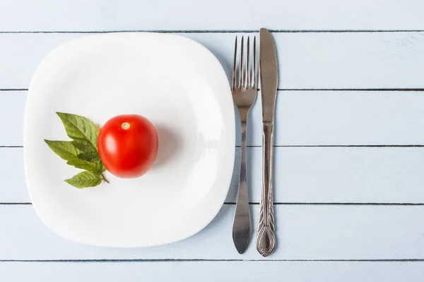 Natural organic tomato with leaves on white plate. Cutlery on wooden table. Vegetarian food concept. Top view with copy space.