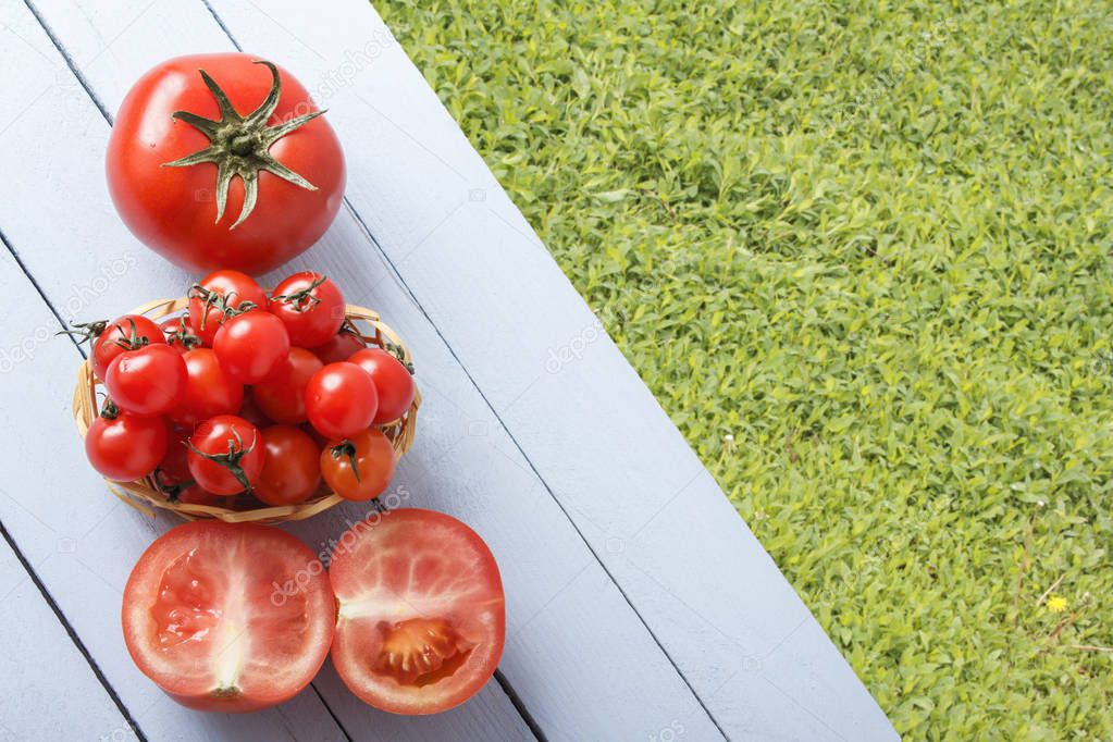 Ripe cherry tomatoes in wicker basket and sliced tomato on wooden table in outdoor. Top view. Grasses for copy space.