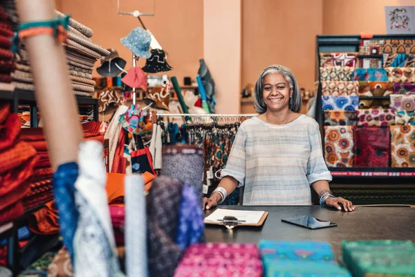 一个微笑成熟的面料店老板的肖像站在她的面料商店柜台后面的彩色布料和纺织品包围 — 图库照片