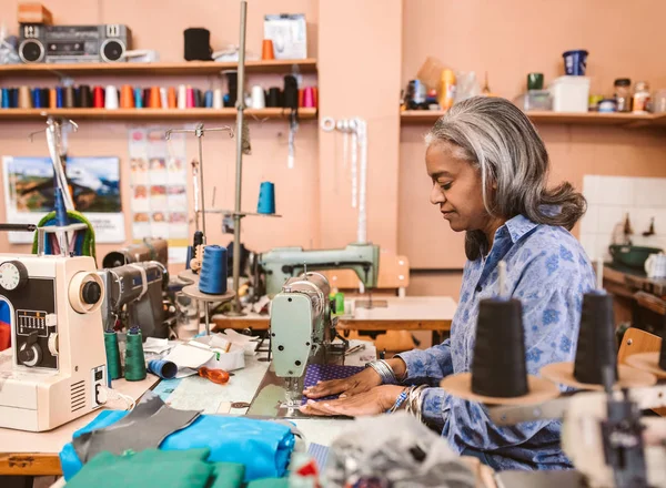 ミシンで作業や刺繍のカラフルな生地の部分彼女の店でテーブルに座っている成熟した女性の裁縫師 — ストック写真