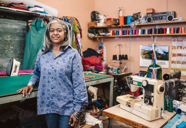 縫製装置とカラフルな布と織物によって彼女の布ワーク ショップ サラウンドのテーブルが立っている成熟した女性の裁縫師の肖像 — ストック写真