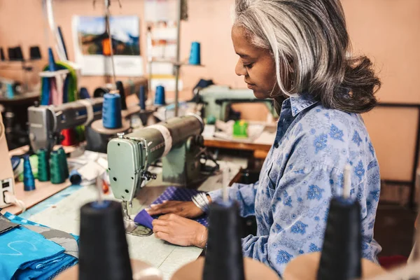 彼女のテキスタイル ショップで一人で仕事をしながらカラフルな生地の部分を縫うミシンを使って成熟した女性の裁縫師 — ストック写真