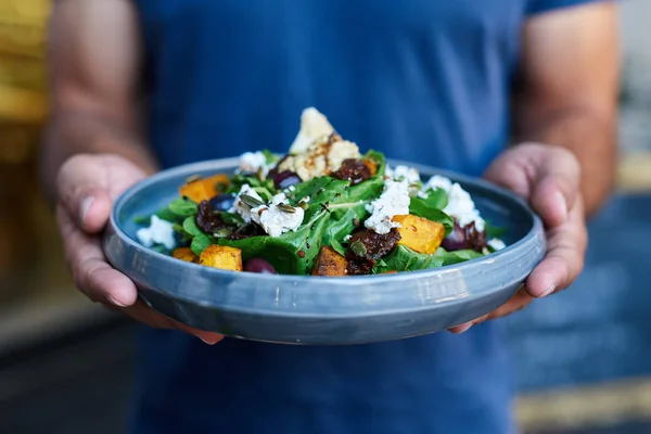 Closeup of a server standing outside of a bistro holding a plate with a delicious mixed salad of butternut, lettuce, olives and feta