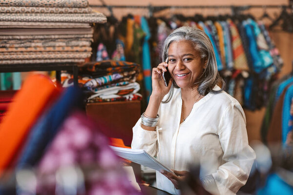 Portrait of a smiling mature fabric shop owner reading paperwork and talking on a cellphone while standing behind a counter surrounded by colorful textiles