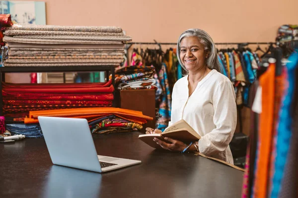 一个微笑成熟的织物商店老板的肖像工作在笔记本电脑上 并写下笔记在一个柜台周围的五颜六色的纺织品 — 图库照片