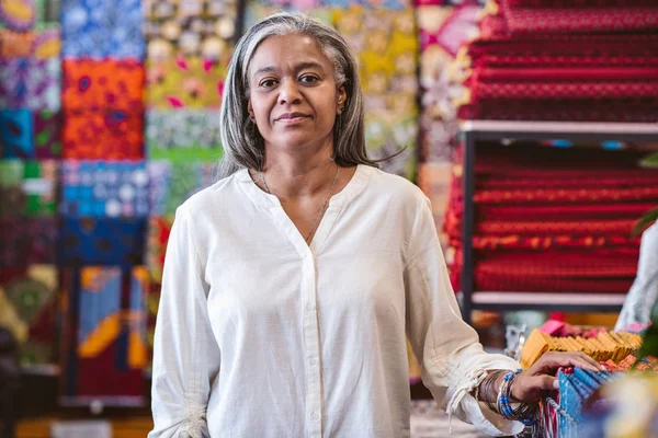 ラックと棚のカラフルな布や繊維の完全に囲まれた彼女の店で立っている成熟した生地店のオーナーの肖像画 — ストック写真