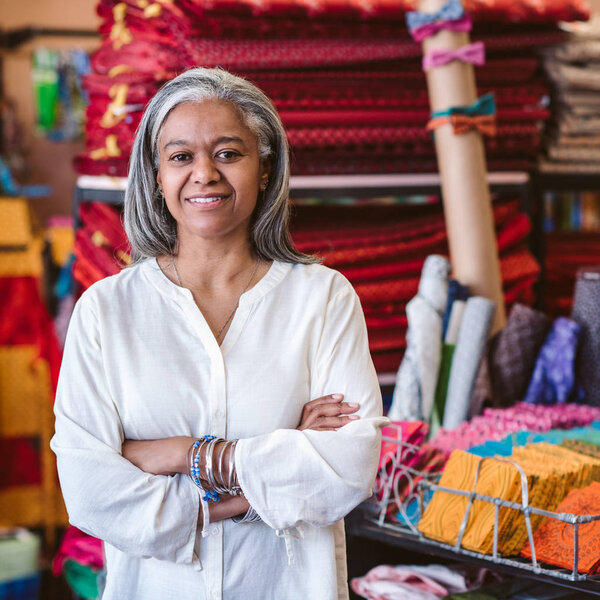 Portrait of a smiling mature fabric store owner standing with her arms crossed by racks and shelves full of colorful cloths and textiles