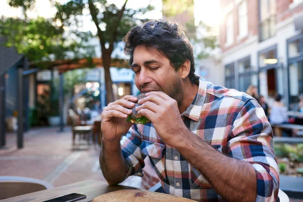 饥饿的年轻人吃了一口他的罂粟籽百吉饼 而坐在他的眼睛封闭在路边咖啡馆表 — 图库照片
