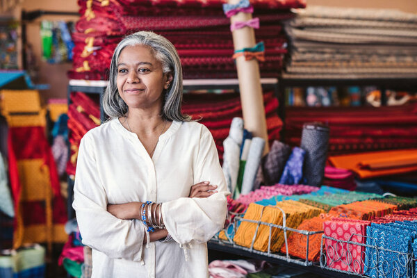 Smiling mature fabric shop owner standing with her arms crossed surrounded by shelves full of colorful textiles and cothes