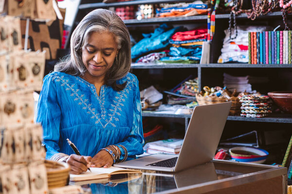 Smiling mature fabric shop owner writing in a notebook and working on a laptop at a counter surrounded by colorful cloths and textiles 