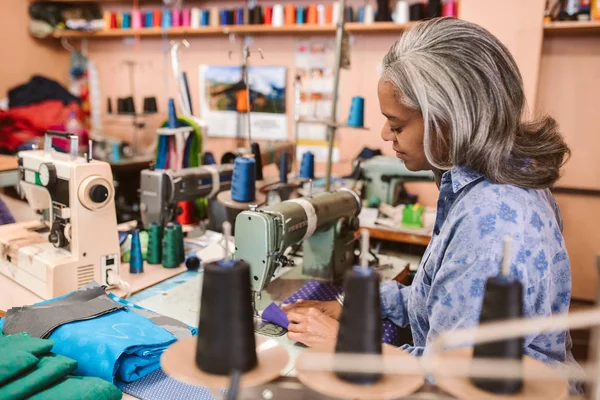 彼女の服のワーク ショップで一人で作業しながらカラフルな生地の部分をステッチ ミシンで座っている成熟した女性の裁縫師 — ストック写真