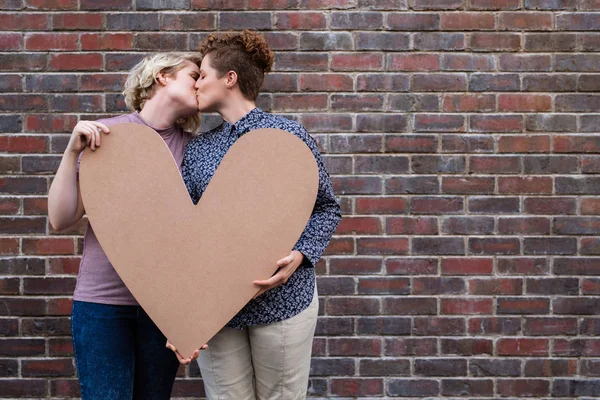 若いレズビアン カップルが外の心のシンボルを保持レンガ壁の前に立っている間キスを愛する — ストック写真