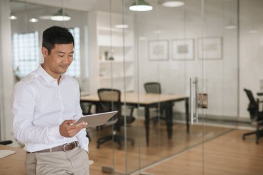 Çevrimiçi bir dijital tabletiyle çalışıyorsanız büyük modern ofisindeki tek başına duran gülümseyen genç Asya iş adamı