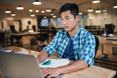 Genç Asyalı tasarımcı online çalışma ile bir dizüstü bilgisayar ve bir not defterinizdeki notlar aşağı geç bir ofiste onun masasında çalışırken yazma