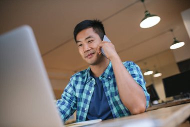 Bir telefonda konuşmak ve bir dizüstü bilgisayar üzerinde çalışan modern bir ofis masasında Asya tasarımcı oturan genç gülümseyen