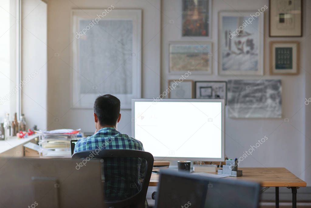 Rear view of a young designer sitting at his desk working on a computer while working late in an office
