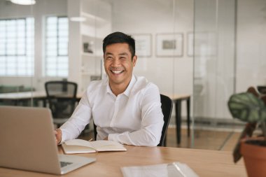 Bir dizüstü bilgisayar ile çalışma ve modern bir ofiste onun masasında otururken notlar aşağı yazma bir gülümseyen Genç Asyalı işadamı portresi