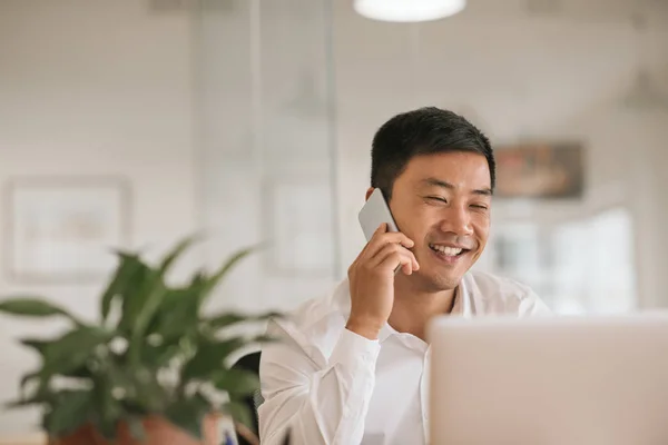 ラップトップに取り組んでいる近代的なオフィスに彼の机に坐っている間彼の携帯電話で話している笑顔の若いアジア系のビジネスマン — ストック写真