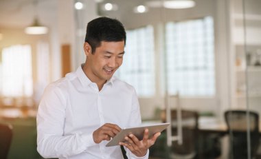 Geniş bir modern ofisi online dijital tablet ile çalışma tek başına ayakta iken gülümseyen genç Asya iş adamı