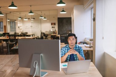 Otururken yalnız modern bir ofiste onun masasında bir odaklı genç Asya tasarımcı çalışma ile bir dizüstü bilgisayar ve monitör portresi