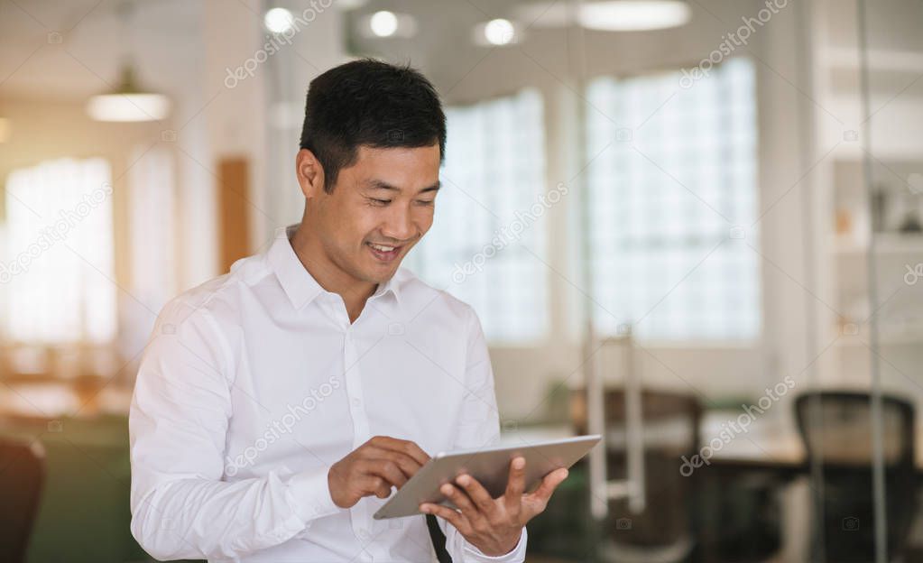 Young Asian businessman smiling while standing alone in a large modern office working online with a digital tablet