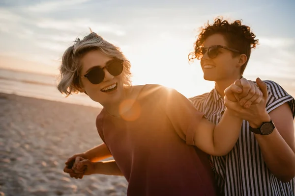 无忧无虑的年轻女同性恋夫妇戴着太阳镜开玩笑地跳舞手一起在沙滩上日落 — 图库照片