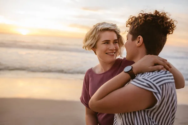夕日を見て砂浜で一緒に若いレズビアンのカップルに立って腕を笑顔 — ストック写真