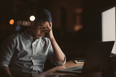 Arka planda şehir ışıkları ile gece geç saatlerde kafasını elinde bir ofiste onun masasında oturan yorgun Asya iş adamı