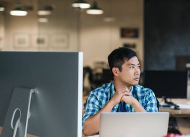 Genç Asyalı tasarımcı düşünme hakkında bir dizüstü bilgisayarda çalışan modern ofisindeki bir masada otururken eserleri