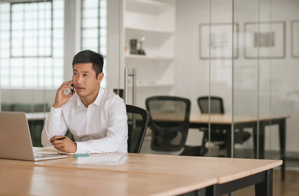 近代的なオフィスに机に彼のラップトップで座りながら会話を彼の携帯電話に若いアジア系のビジネスマン — ストック写真