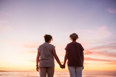 Dikiz içerik genç lezbiyen çift ayakta el el birlikte romantik bir gün batımını izlemek okyanus üzerinde gökyüzü karşı