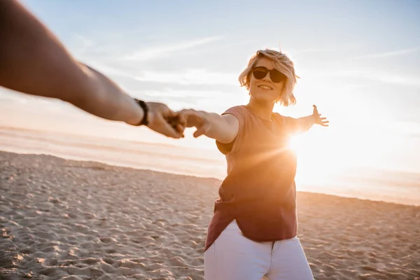 微笑着戴着墨镜的年轻女子手持她的女朋友的手 而带领她沿着沙滩在日落 — 图库照片