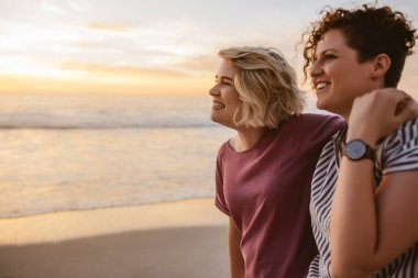 Genç lezbiyen çift ayakta kol kola okyanus üzerinde gün batımını izlemek birlikte bir kum plajındaki gülümseyen