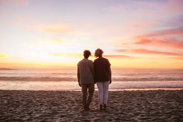 一对年轻的女同性恋夫妇站在一起在沙滩上观看浪漫的日落在海洋的后方视图 — 图库照片