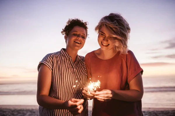 两个年轻的女朋友笑和玩烟花 同时在海滩上的乐趣在黄昏 — 图库照片