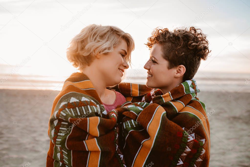 Smiling young lesbian couple wrapped up together in a warm blanket while enjoying sunset at the beach