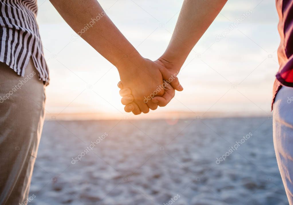 Cropped closeup of a lesbian couple holding hands while standing on a sandy beach watching the sunset together 