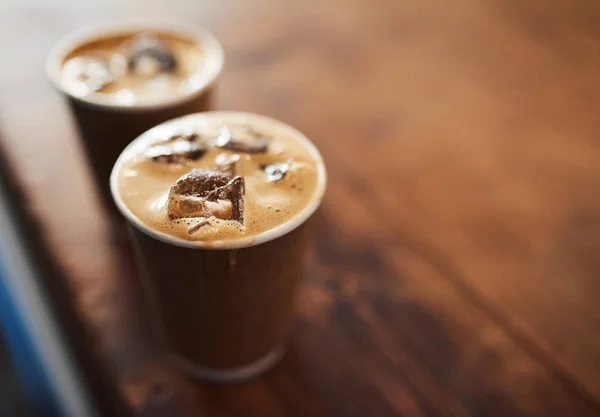 Closeup of two ice coffees in takeaway cups sitting next to each other on the wooden counter of a cafe