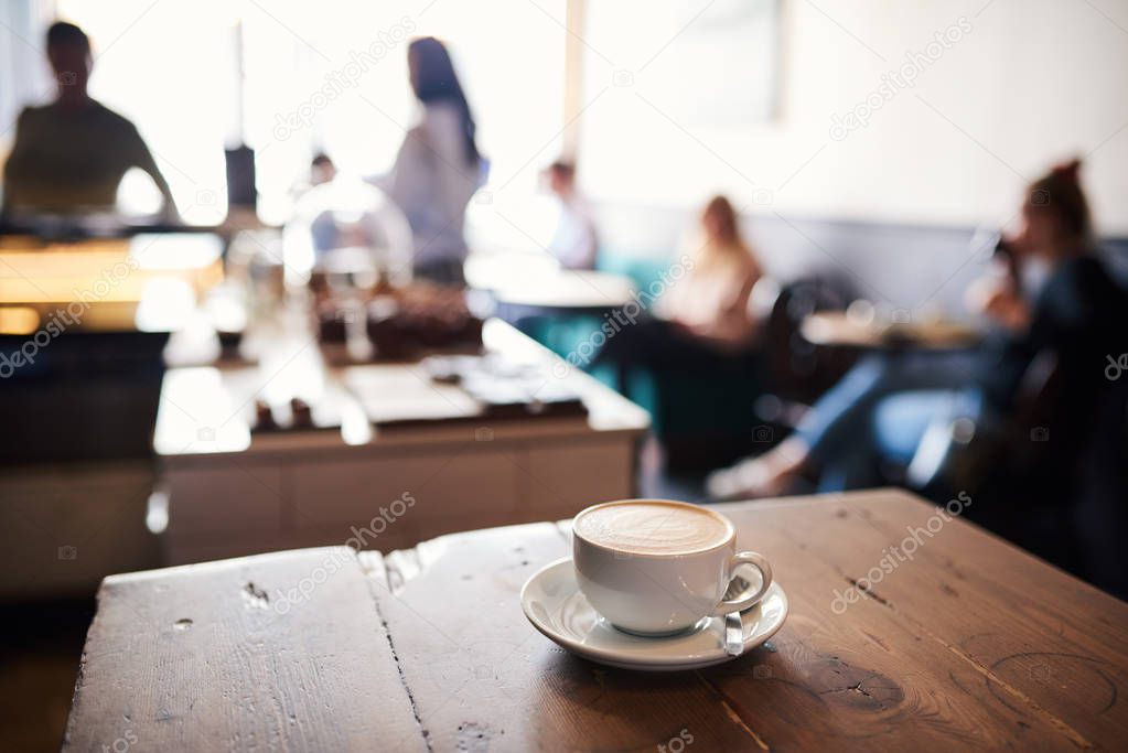 Cup of cappuccino sitting on a wooden counter in a trendy cafe with customers in the background