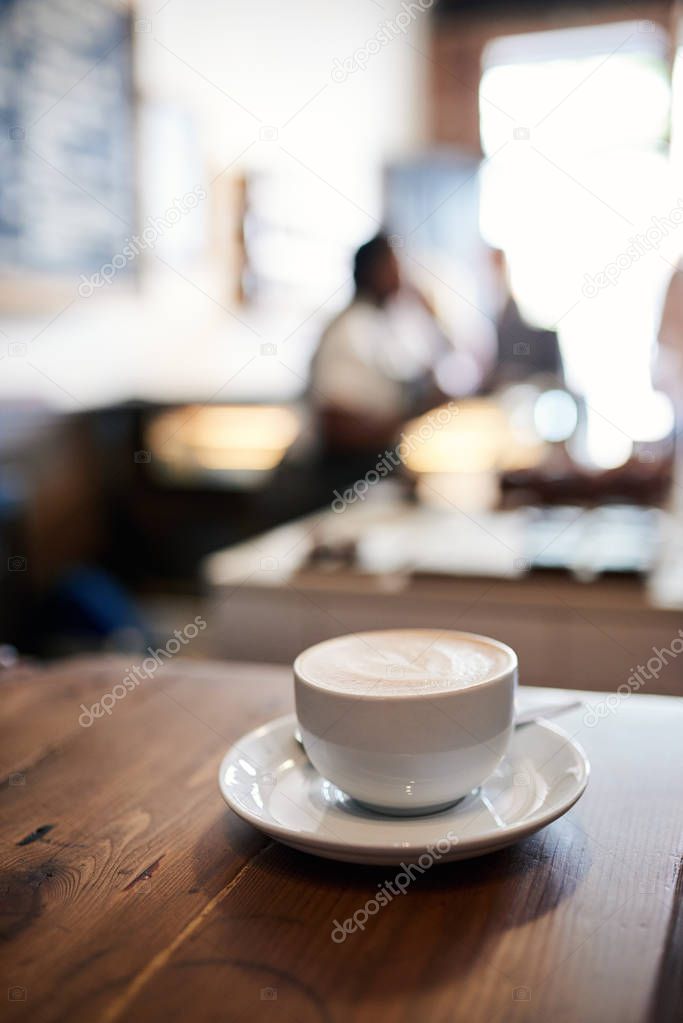 Cup of cappuccino with a heart shape in the foam sitting on a counter in a cafe with customers in the background