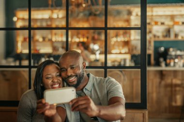 Smiling young African couple taking a selfie together while sitting a table in a trendy bar clipart