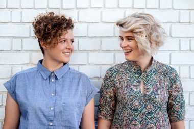 Genç lezbiyen çift gülümseyen ve birlikte bir tuğla duvarın dışında önünde ayakta iken birbirine bakarak